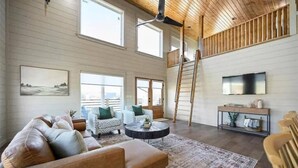 Un salón amplio con techo alto, ventanas grandes, chimenea y televisor montado en la pared.