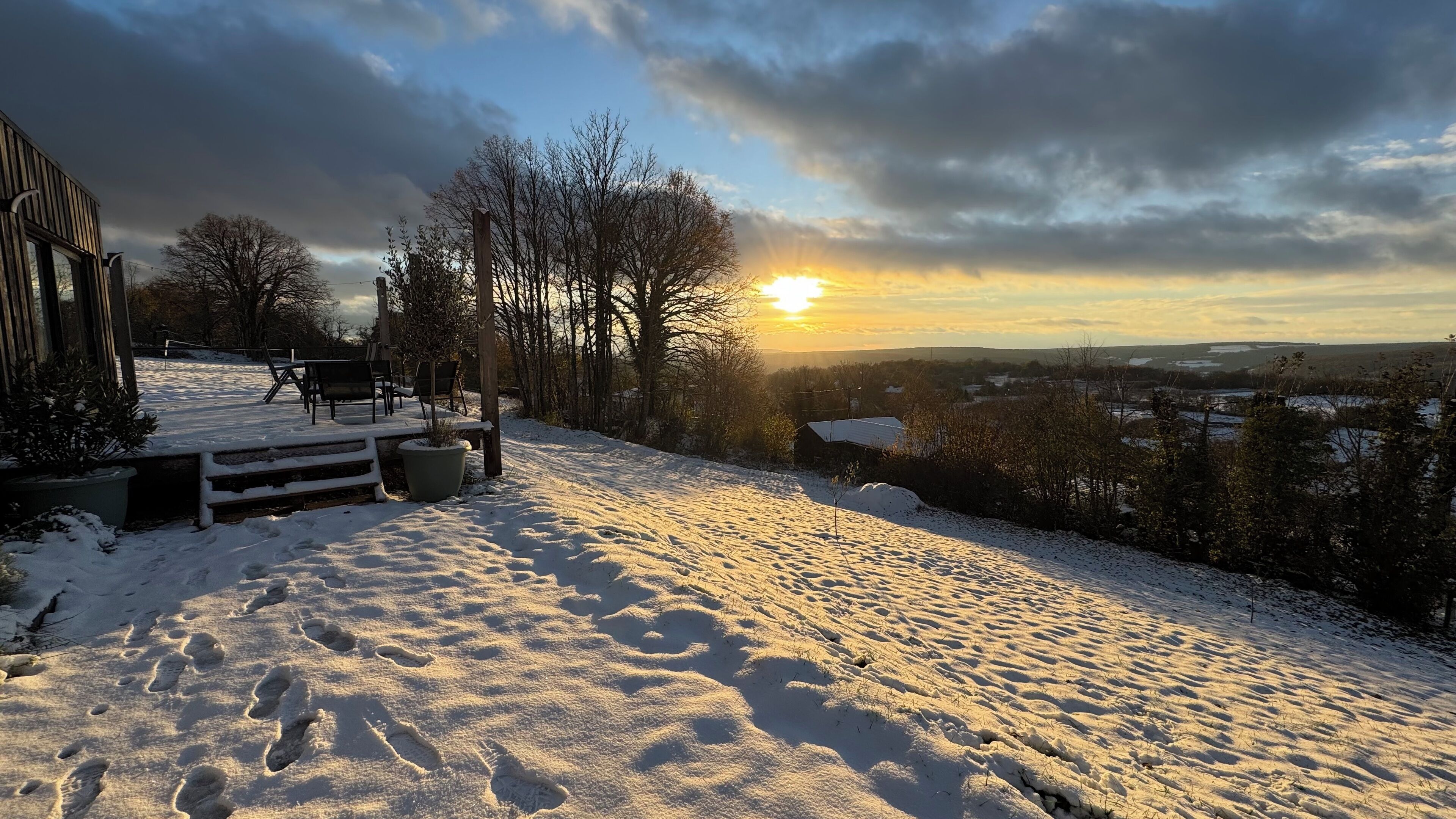 A snow-covered deck with outdoor seating, a view of the countryside, and a sunset in the background.