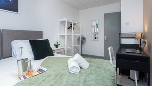 A hotel room with a bed, a desk with a laptop, a mirror, a wardrobe, and a fan.