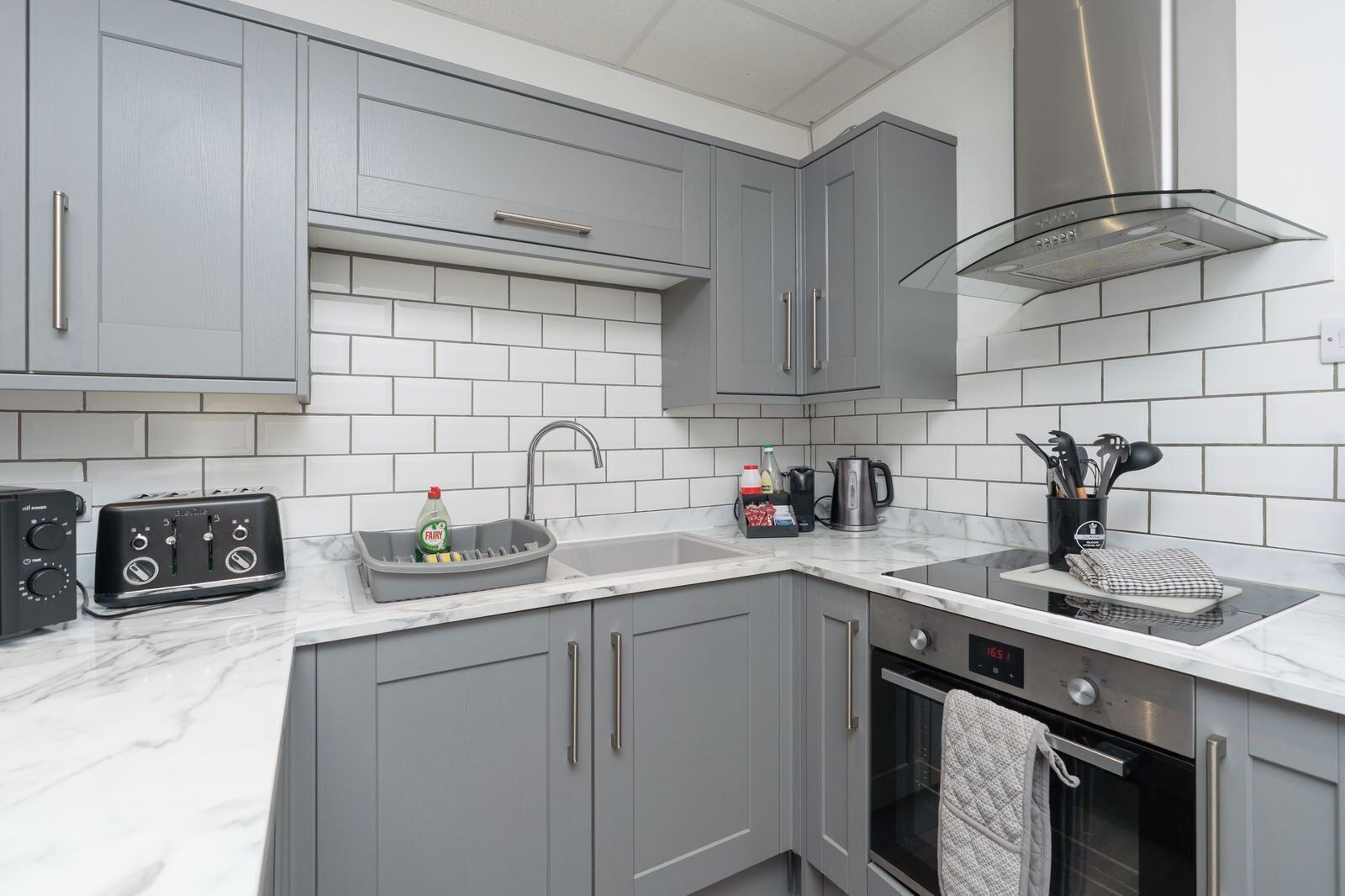 A kitchen with grey cabinets, a stainless steel range hood, a sink with a dish rack, a toaster, and a kettle.
