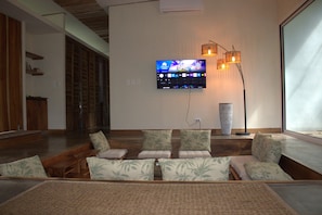A modern living room with a flat-screen TV mounted on the wall, a large window, and a seating area with patterned cushions. - Casa Bromelia - A Floating Jungle Retreat - Pets Allowed (Puntarenas)