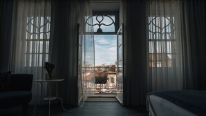 Een kamer met een bed, een klein tafeltje met een plant en uitzicht op een balkon met een hekwerk en gebouwen op de achtergrond.