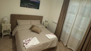 A neatly made bed with a headboard, a bedside table with a lamp, a wall-mounted picture, and a window with curtains. - Casa Marta in Lucca within walking distance of downtown and hospital, private parking (Lucca)