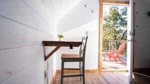 Ein Zimmer mit weißen Holzwänden, einer Holzboden, einer Holzdecke, einem grünen Stuhl, einer hölzernen Bank, einer kleinen Topfpflanze und einer Tür, die zu einem Außenbereich mit einem roten Stuhl führt.