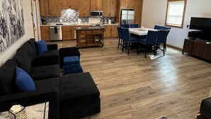 Ein großzügiges Wohnzimmer mit einem schwarzen Ecksofa, einem Essbereich mit blauen Stühlen, einer Küche mit Holzschränken und einem großen Fernseher.