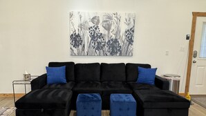 Ein schwarzes Ecksofa mit blauen Kissen und passenden Hocker, ein abstraktes Gemälde in Grau an der Wand und eine Holztür mit Fenster.