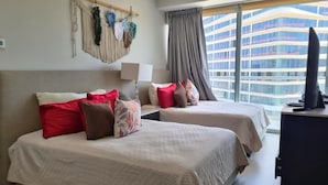 A hotel room with two beds, a desk, a TV, and a large window with a view of a modern building. - Luxury apartment on the beach with beautiful views (Puerto Vallarta)