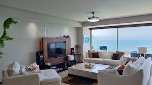 A modern living room with a large flat-screen TV, two white sofas, a coffee table, and a large window offering a view of the sea. - Luxury apartment on the beach with beautiful views (Puerto Vallarta)
