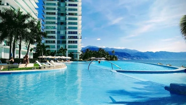 A swimming pool with a view of mountains and a tall building in the background. - Luxury apartment on the beach with beautiful views (Puerto Vallarta)