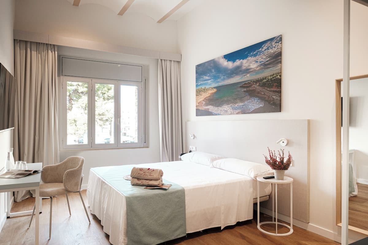 A bedroom with a bed, a desk, a chair, a window with curtains, a painting, and a small table with a plant.