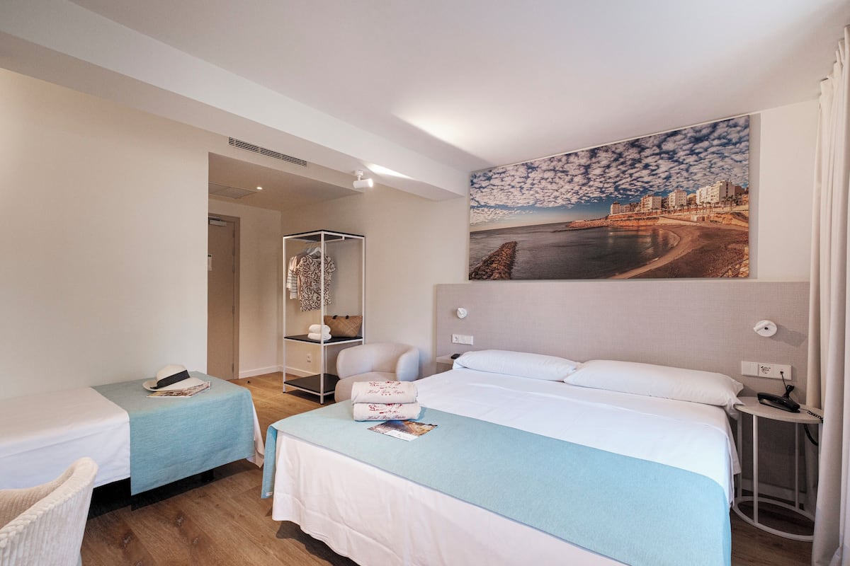 A hotel room with two beds, a large window with a cityscape view, a bedside table with a lamp, and a wardrobe.