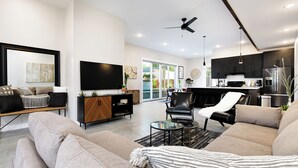 Uma sala de estar moderna com um sofá, uma mesa de centro e uma televisão. Uma cozinha visível ao fundo possui armários escuros e uma geladeira.