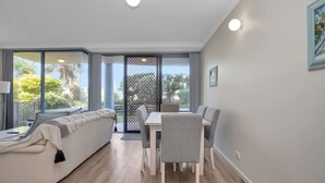 A modern living room with a sofa, a dining table with chairs, and a large sliding glass door leading to an outdoor area. - Nautilus 2 Ground Floor Apartment (Hawks Nest)