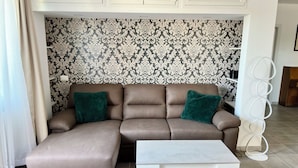 A living room with a brown sectional sofa, two green cushions, a white coffee table, and a patterned wallpaper background. - C Room with shared bathroom; Sweet Home Fleming (Roma)