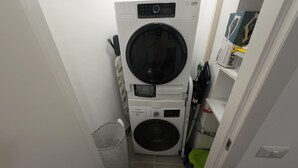 A compact laundry room with a front-loading washer and dryer stacked on top of each other, a small trash bin, and a shelf with a box and other items. - C Room with shared bathroom; Sweet Home Fleming (Roma)