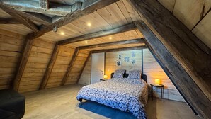 Ein gemütliches Schlafzimmer im Dachgeschoss mit schrägen Holzwänden, einem großen Bett mit gemusterten Betttextilien und einem kleinen Nachttisch mit Lampe.
