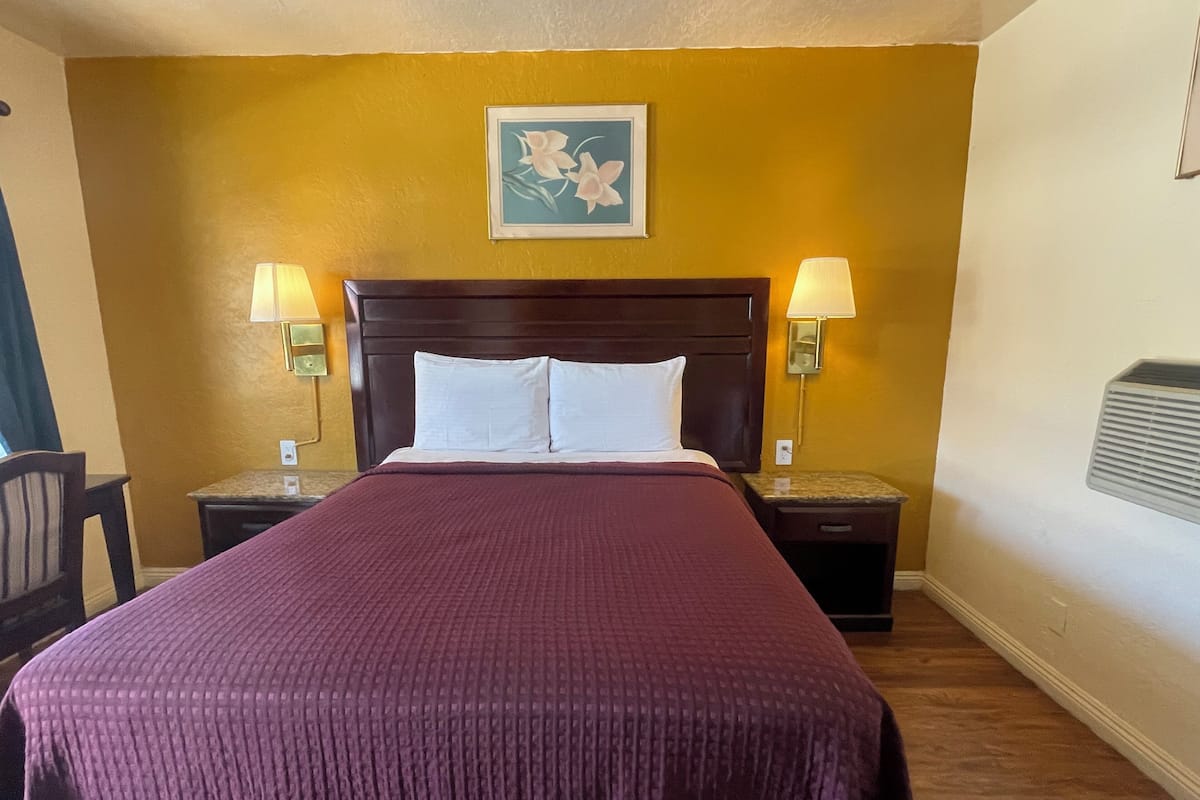 Ein Hotelzimmer mit einem großen Bett, zwei Nachttischlampen, einem Nachttisch, einem gerahmten Blumenbild an der Wand und einer Wand-Klimaanlage.