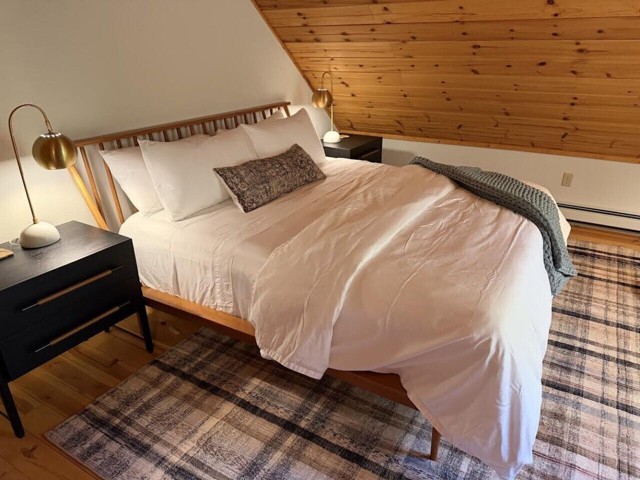 A bedroom with a wooden ceiling, a bed with white bedding, a black nightstand, a lamp, and a plaid rug.
