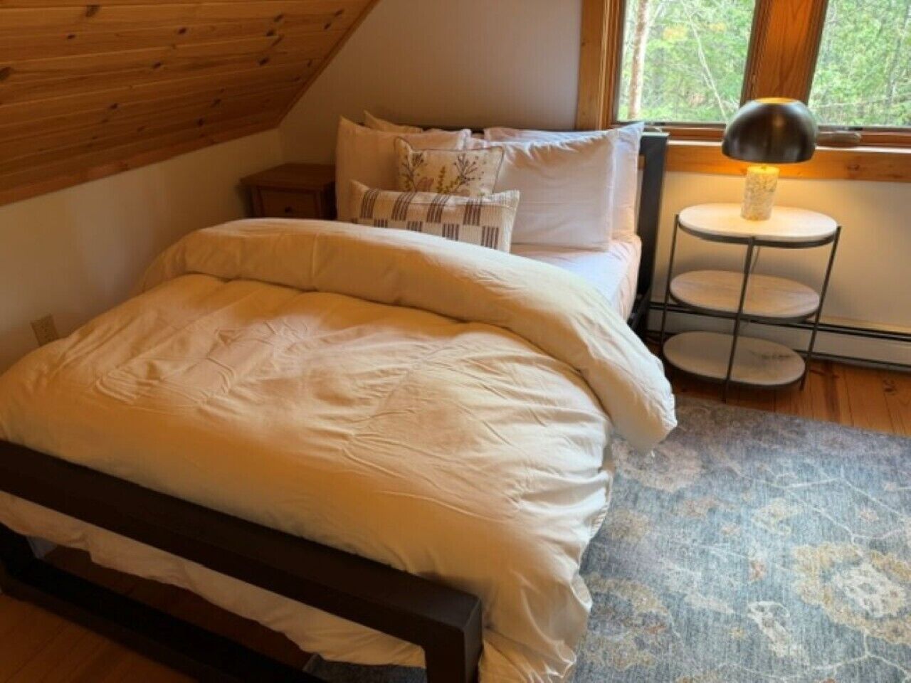 A bedroom with a wooden ceiling, a bed with white bedding, a small bedside table with a lamp, and a view of trees through a window.