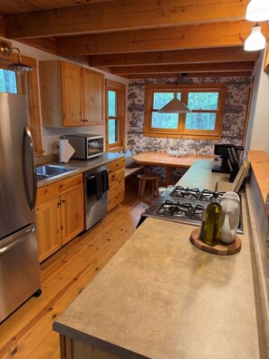 A kitchen with wooden cabinets, a stainless steel refrigerator, a microwave, a gas stove, and a window with a view of trees.