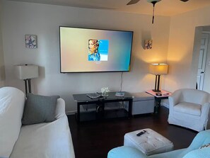 Ein Wohnzimmer mit einem an der Wand montierten Flachbildfernseher, einem kleinen Tisch mit Lampe, einem Sofa und einem Hocker.
