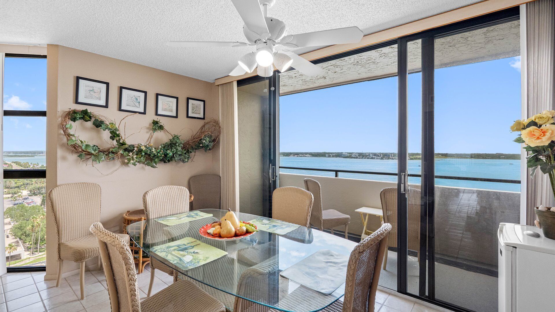 Un balcon avec une table en verre, des chaises en osier, un ventilateur de plafond, des tableaux encadrés et une vue sur la mer.