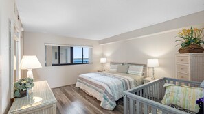 A bedroom with a bed, bedside tables, a crib, a window with a view, and a dresser. - Lighthouse Towers - 1808 (Clearwater)