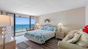 A bedroom with a bed, a nightstand, a lamp, a chair, a sofa, a balcony with a view of the sea, and a decorative wall piece. - Lighthouse Towers - 1808 (Clearwater)
