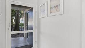Ein Flur mit einer Glastür, die zu einem Außenbereich mit Palmen und einem Gebäude im Hintergrund führt. An der Wand hängen zwei gerahmte Kunstwerke.