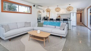 A modern living room with a sofa, coffee table, and a kitchen area in the background. - Summer Palms - Tairua Holiday Home (Tairua)