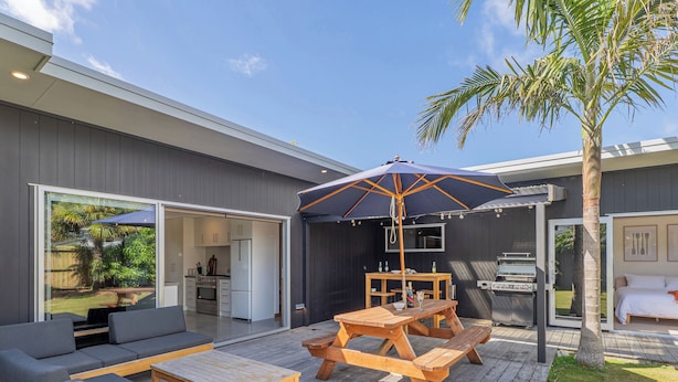 An outdoor patio with a wooden table and benches, a grill, and a large umbrella. - Summer Palms - Tairua Holiday Home (Tairua)