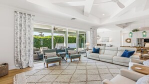 Ein geräumiges Wohnzimmer mit einer großen Couch, zwei Sesseln und einem Couchtisch. Eine Glastür führt auf eine Terrasse mit Gartenmöbeln.
