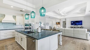 Eine moderne Küche mit Kochinsel, weißen Schränken und einer blauen Arbeitsplatte. Es gibt drei hängende Pendelleuchten, einen eingebauten Backofen und ein großes Flachbildfernsehen im Wohnbereich.