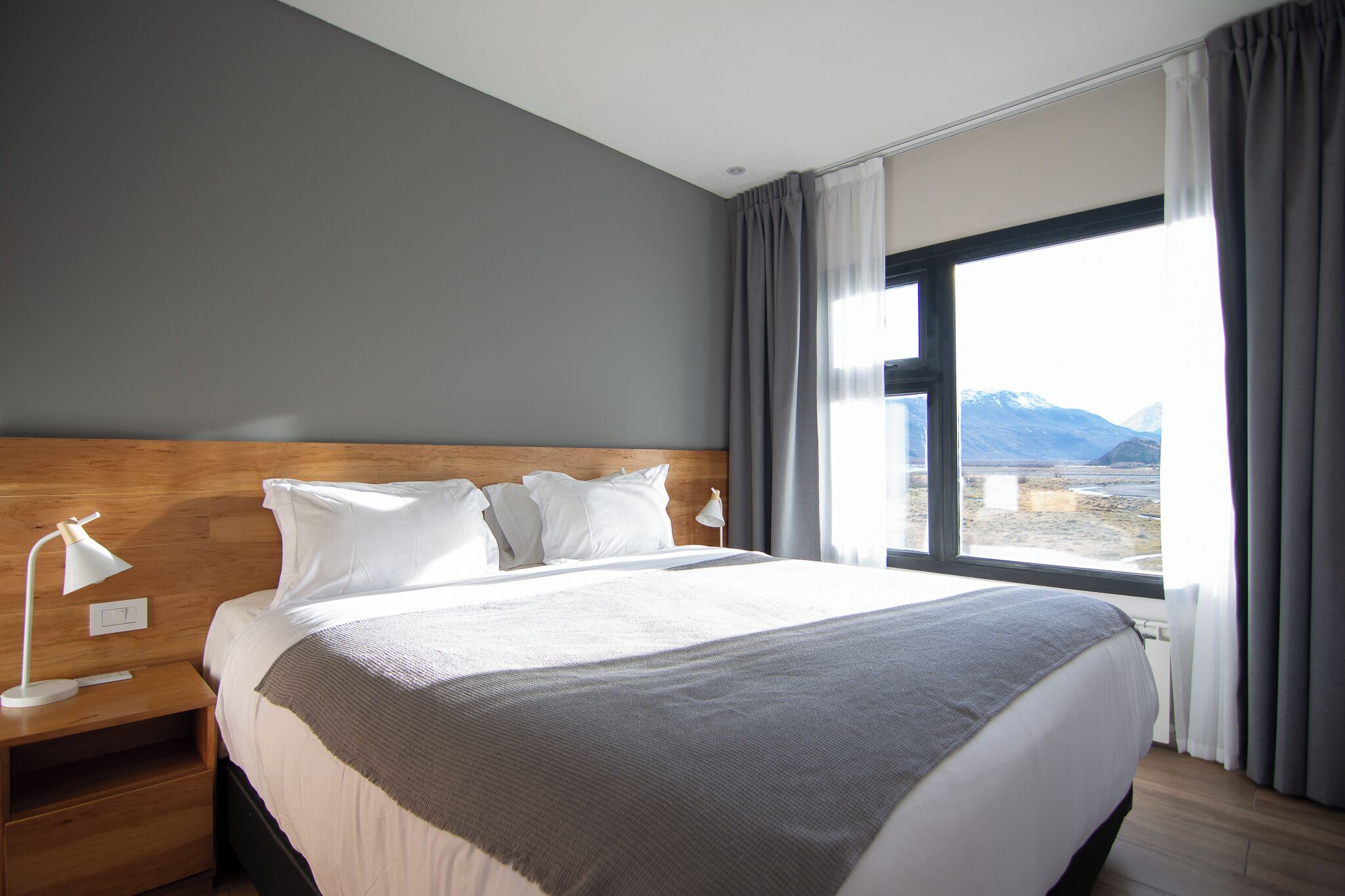 Habitación de hotel con una cama grande, una mesita de noche, una lámpara y una ventana con vistas a la montaña.