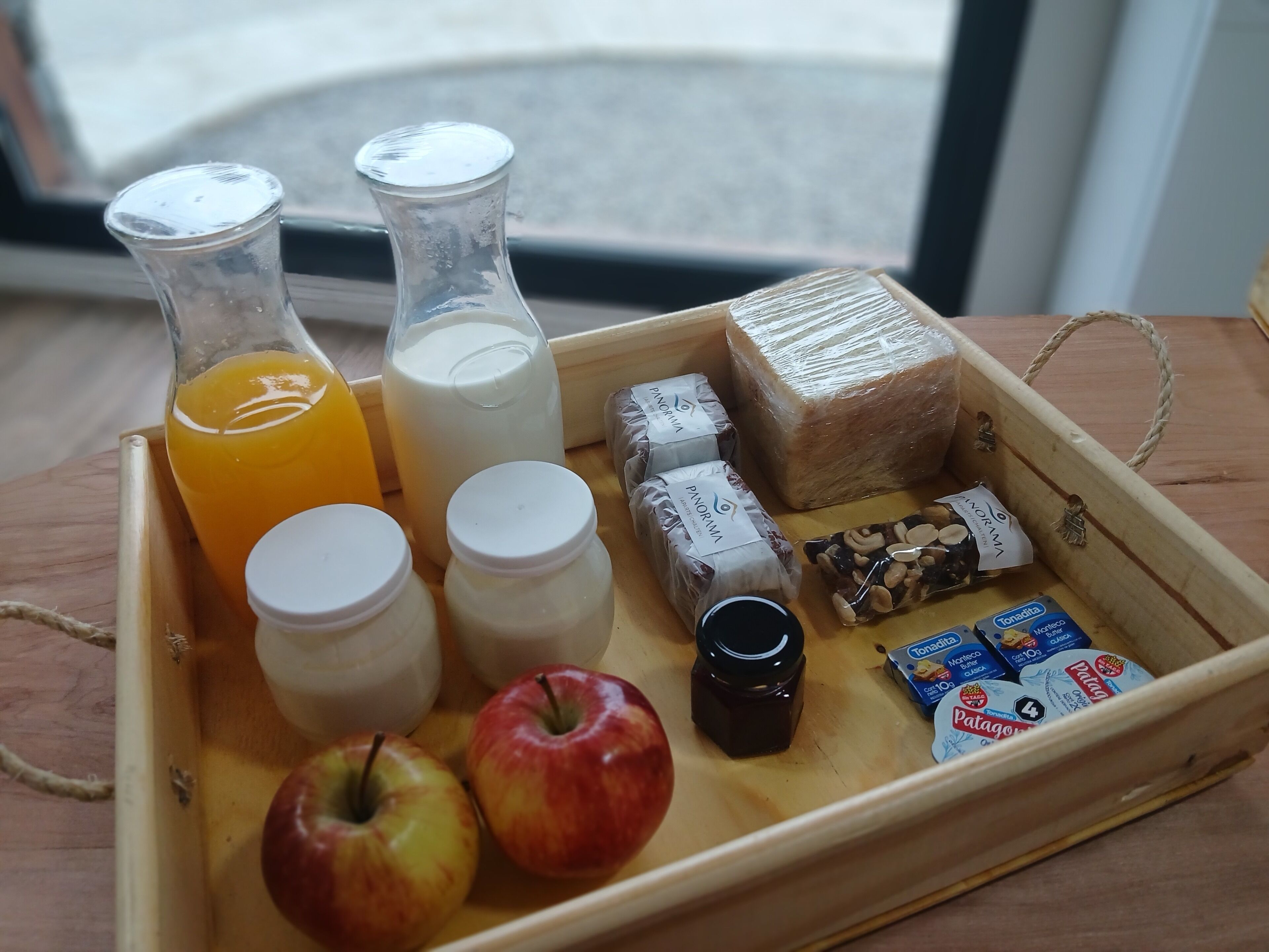 Una bandeja de madera con una botella de vidrio de jugo de naranja, una botella de vidrio de leche, dos frascos, dos manzanas, un frasco pequeño de mermelada, una pastilla de jabón y una caja de galletas Patagónicas.