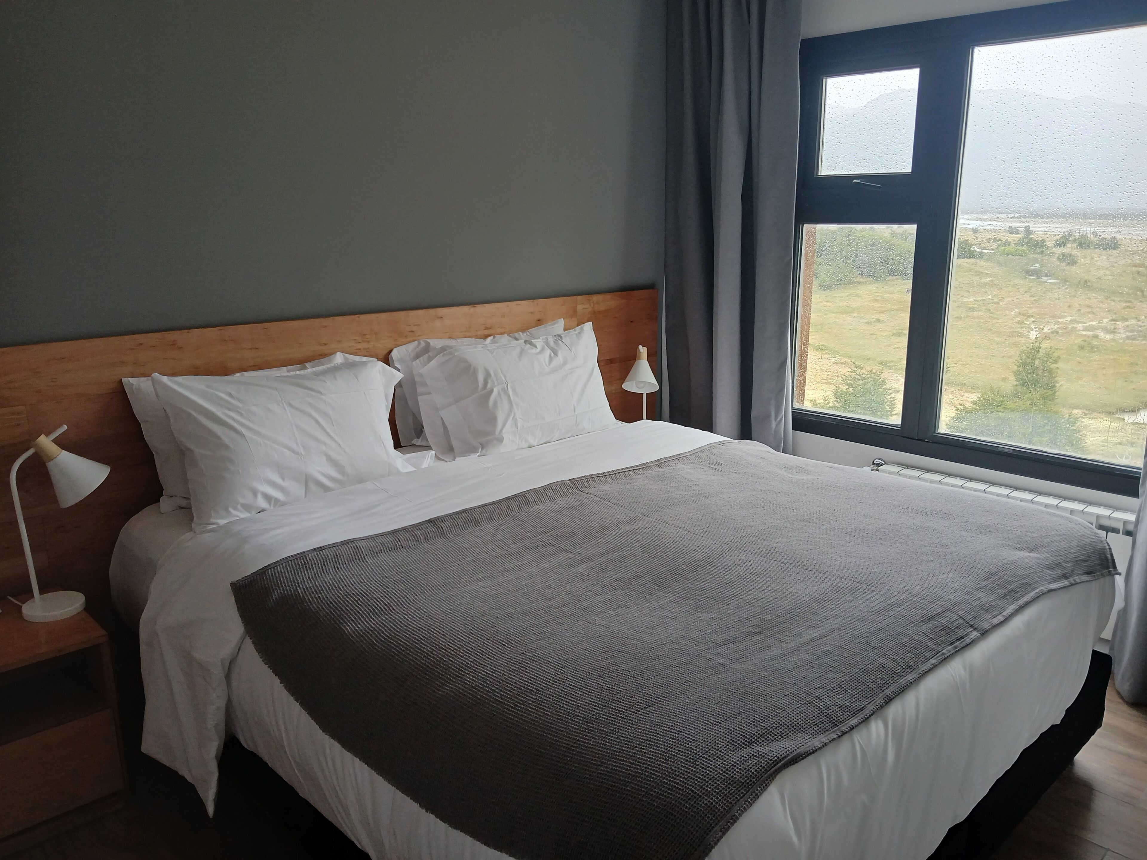 Een netjes opgemaakt bed met witte lakens en een grijze deken, een houten hoofdbord, een bedlampje en een raam met uitzicht op een landschap.