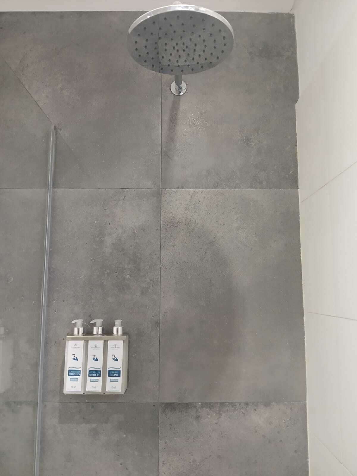 A shower with a round head, grey tiles, and three bottles of shampoo on the wall.