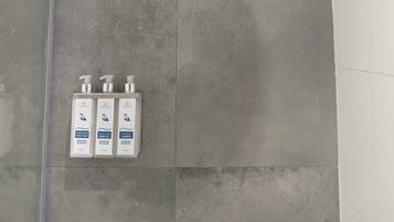A shower with a round head, grey tiles, and three bottles of shampoo on the wall.