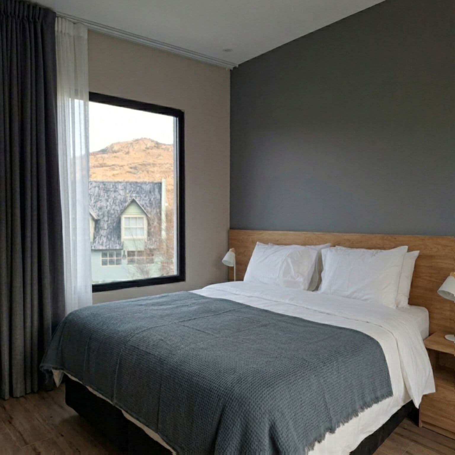 A bedroom with a large window, a bed with white linens, a wooden headboard, and a grey blanket.