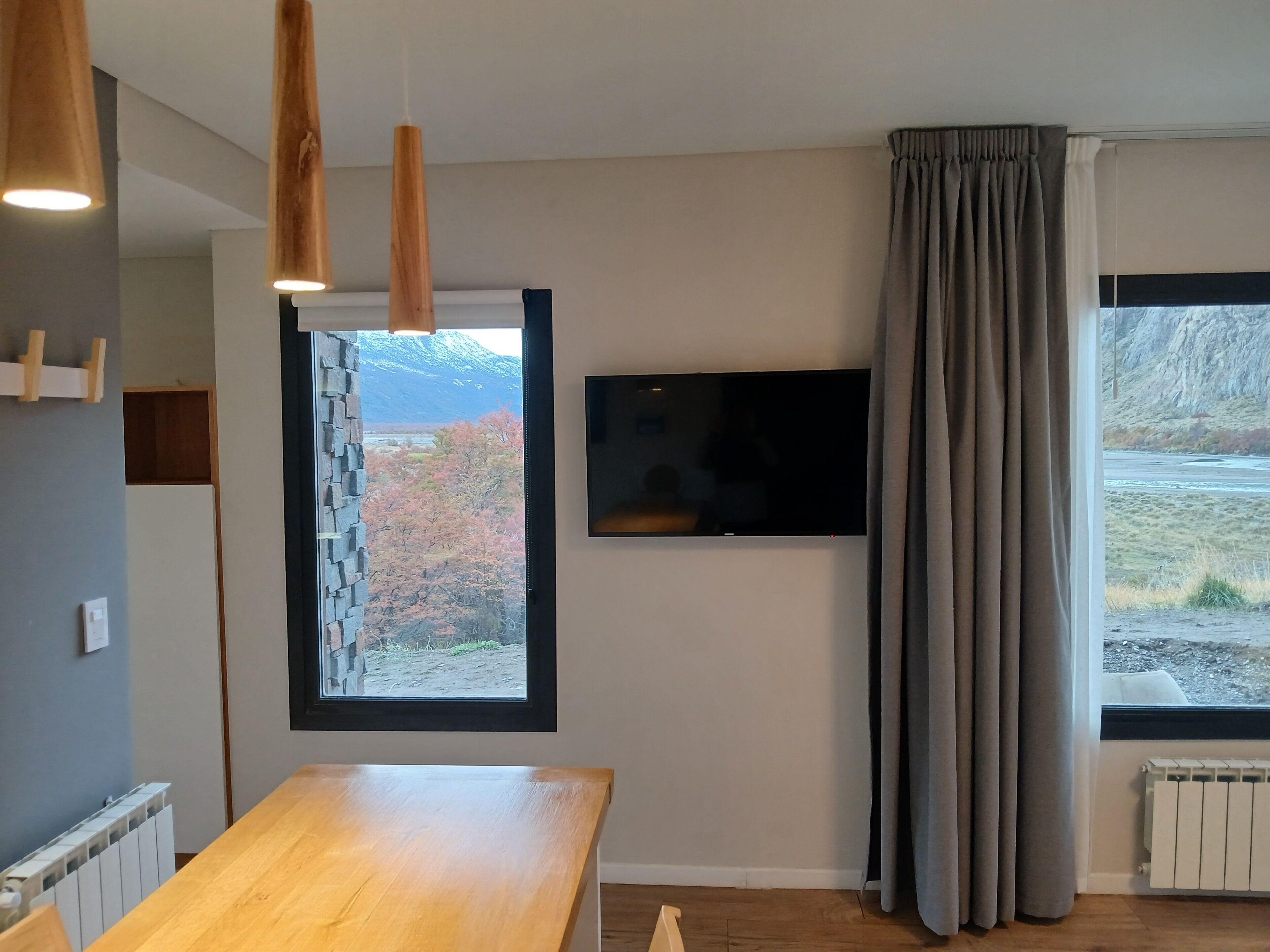 A modern room with a wooden dining table, a mounted TV, a stone wall, and a window with curtains.