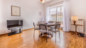 A modern living room with a flat-screen TV, a dining table with chairs, a lamp, and a large window with curtains. - Conca dóro - Val Dála Retreat (Rome)
