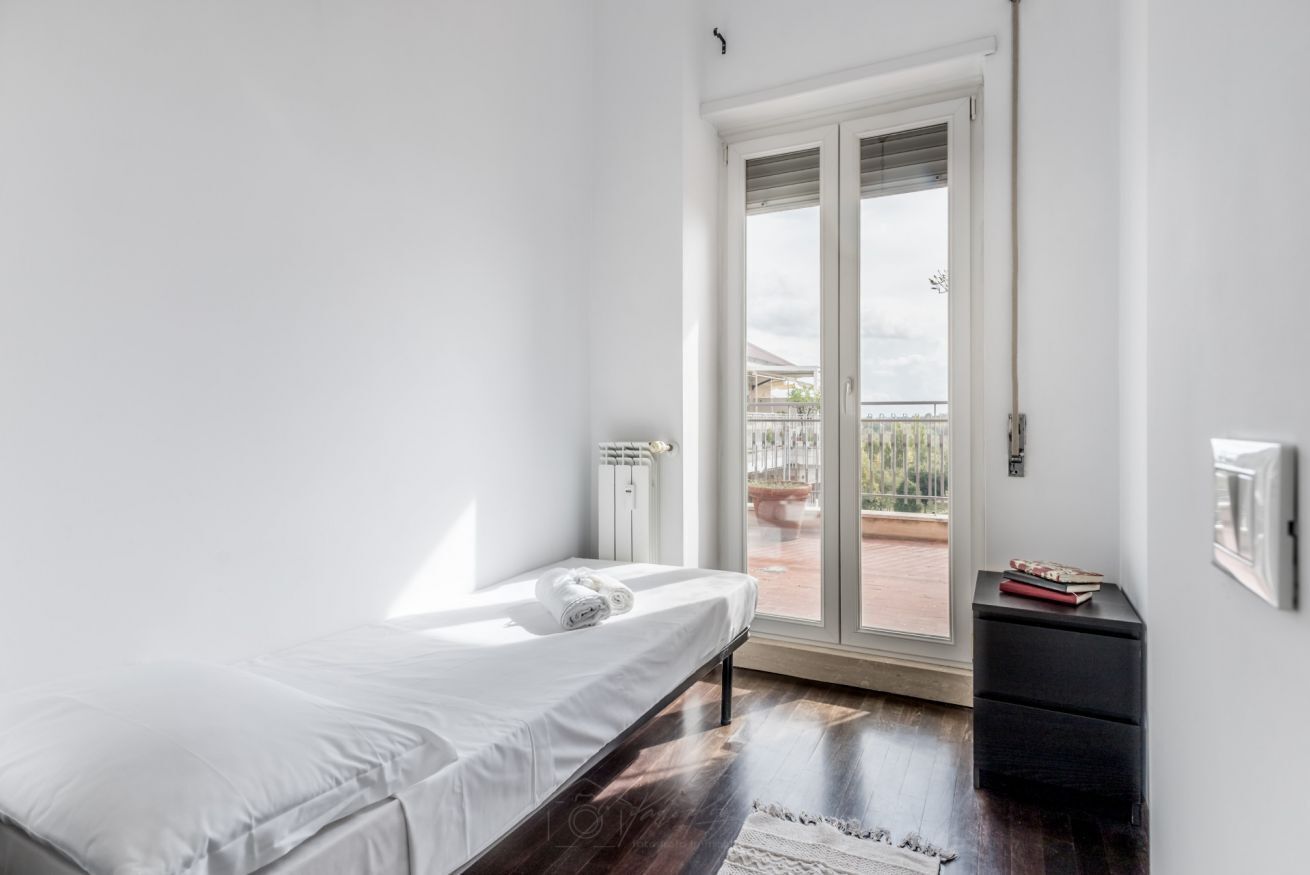 Camera da letto minimalista con un letto singolo, un comodino nero e vista su un balcone.