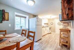 A compact kitchen with a refrigerator, a sink, and wooden cabinets. There is a dining table with chairs and a window with a view of trees.