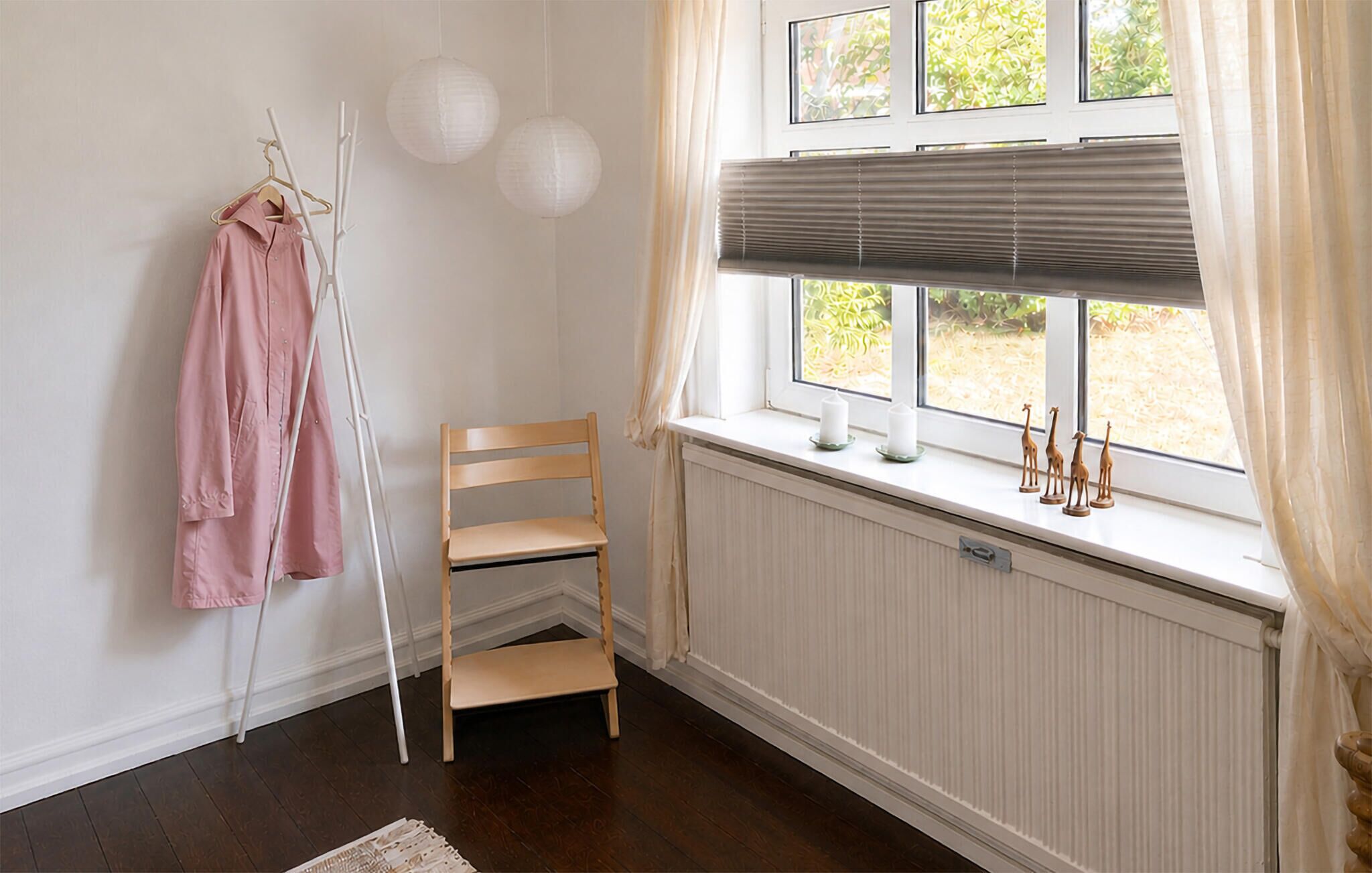 Una habitación con un radiador, una ventana con persianas, una silla, un perchero con una prenda rosa y un pequeño estante con adornos.