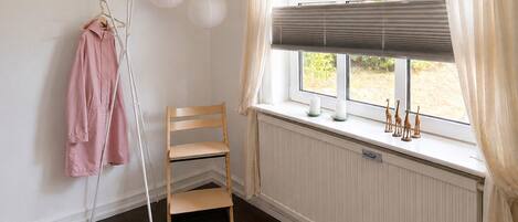 Una habitación con un radiador, una ventana con persianas, una silla, un perchero con una prenda rosa y un pequeño estante con adornos.