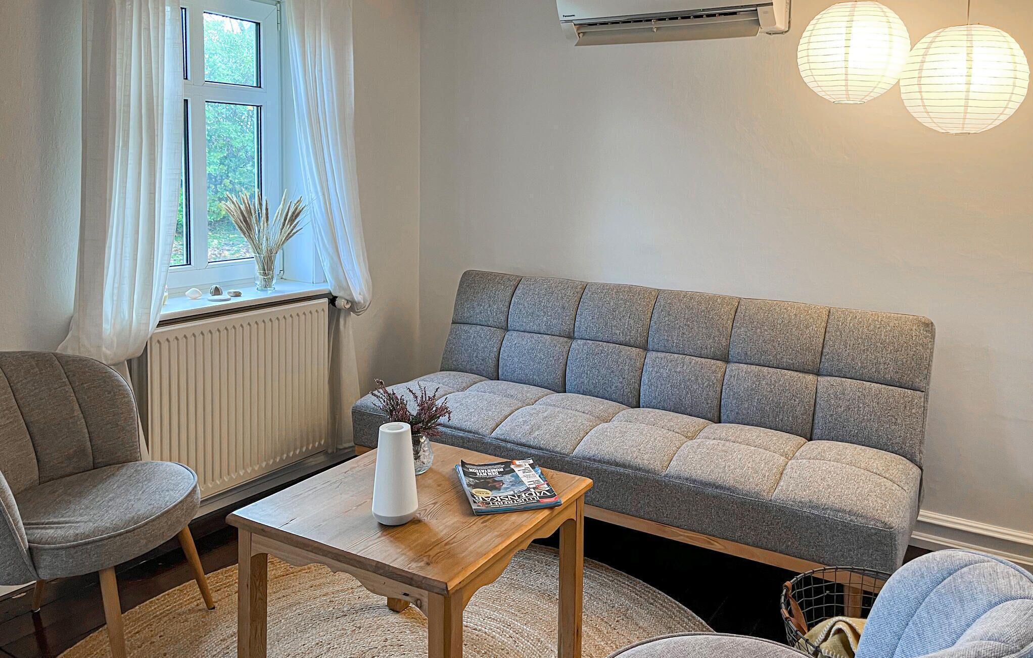 Een gezellige woonkamer met een grijze bank, een houten salontafel en een raam met witte gordijnen.