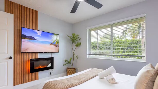A bedroom with a bed, a TV mounted on a wooden panel wall, a fireplace, a window with blinds, and a ceiling fan.