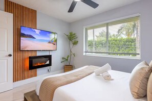 A bedroom with a bed, a TV mounted on a wooden panel wall, a fireplace, a window with blinds, and a ceiling fan. - Stylish 3BR Haven — Minutes to Beach & Downtown FTL (Fort Lauderdale)