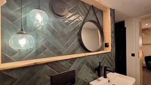 Ein Badezimmer mit Fischgrätmuster-Fliesen an der Wand, einem spiegel mit Holzrahmen, einem weißen Waschbecken und Pendelleuchten.