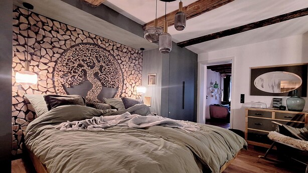 Ein Schlafzimmer mit Steinwand, einem Bett mit grüner Decke, einem hölzernen Schminktisch und einer Pendelleuchte.